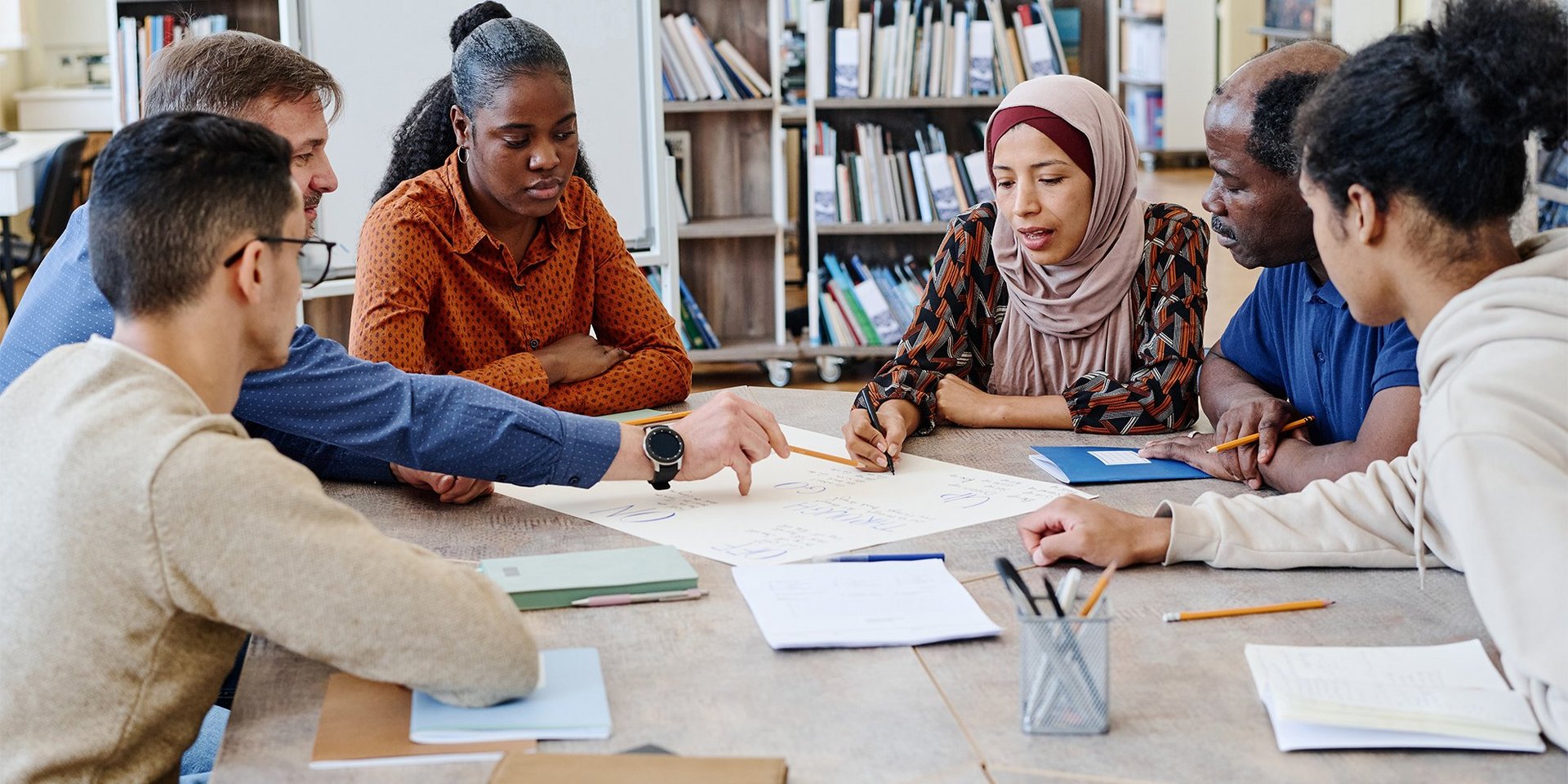 Eine Gruppe Internationals mit Lehrkraft sitzt an einem Tisch in einer Bibliothek und arbeitet an einer Aufgabe. 