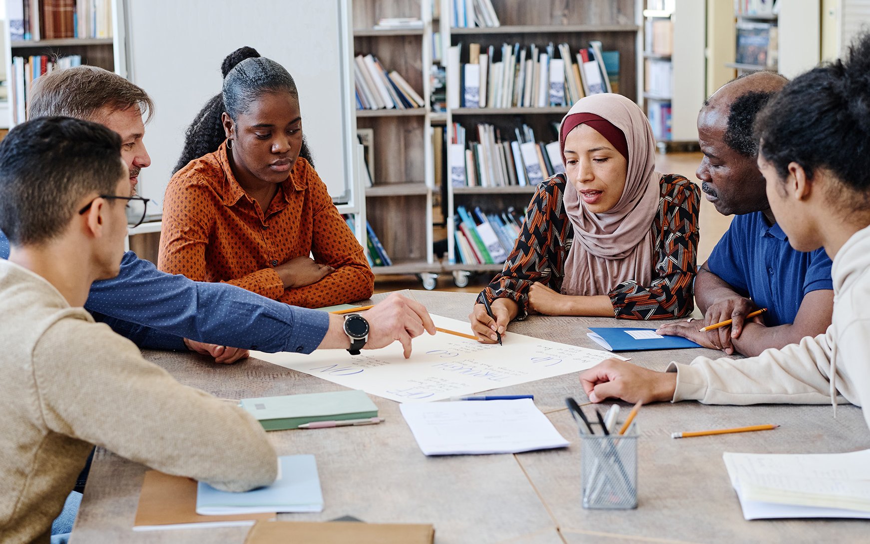 Deutsch-Test für Zuwanderer | AdobeStock Eine Gruppe Internationals mit Lehrkraft sitzt an einem Tisch in einer Bibliothek und arbeitet an einer Aufgabe.