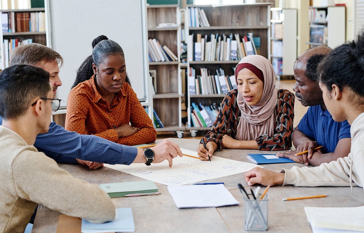 Eine Gruppe Internationals mit Lehrkraft sitzt an einem Tisch in einer Bibliothek und arbeitet an einer Aufgabe. 