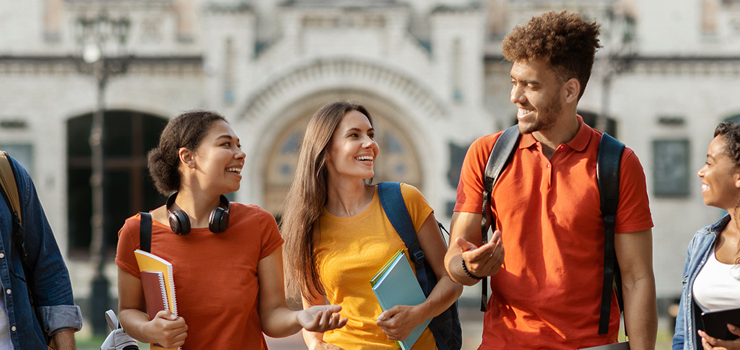 Fünf Studierende laufen mit Rucksack und Studienmaterialien vor einem historischen Universitätsgebäude entlang.