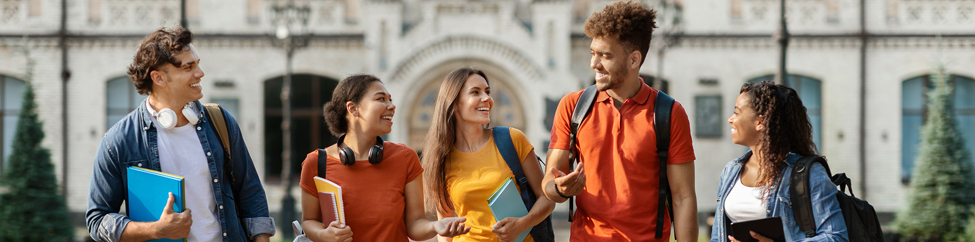 Fünf Studierende laufen mit Rucksack und Studienmaterialien vor einem historischen Universitätsgebäude entlang.
