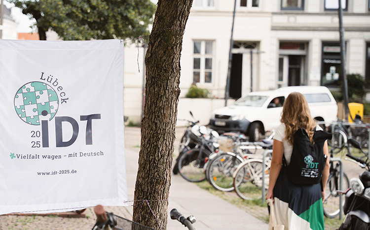 „Vielfalt wagen – mit Deutsch“, das Motto der IDT | Foto: IDT Banner der IDT mit dem Motto „Vielfalt wagen – mit Deutsch“ gespannt zwischen zwei Bäumen, auf der rechten Seite ist eine Teilnehmerin in Rückenansicht zu sehen, die einen Veranstaltungsrucksack trägt.